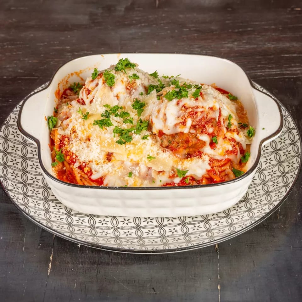 Baked pasta with melted cheese, red sauce, and fresh parsley in a white dish on a patterned plate.