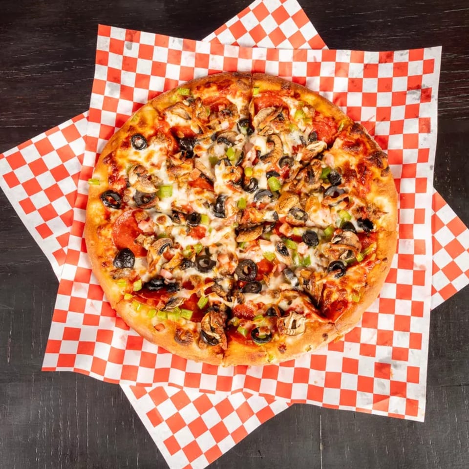 A whole pizza with pepperoni, mushrooms, olives, and green peppers on red and white checkered paper.