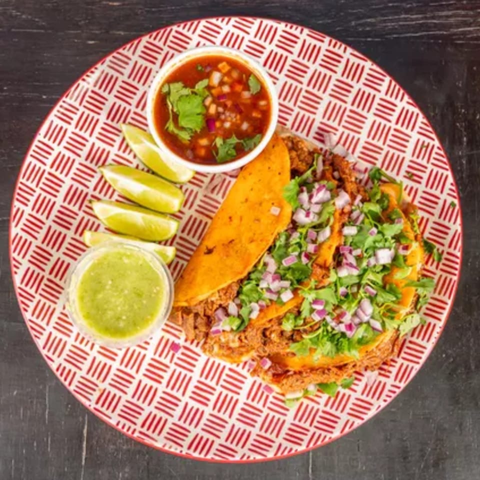 Two birria tacos with cilantro and red onions, served with consome, salsa verde, and lime wedges.